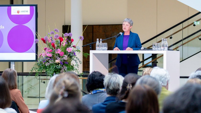 Ansicht der Portikushalle während der Veranstaltung. Im Vordergrund sind einige Hinterköpfe des Publikums zu sehen. Gegenüber auf der Bühne steht Landtagspräsidentin Hanna Naber am Redepult. Links neben ihr steht ein großer Bildschirm mit dem Titel der Veranstaltung.