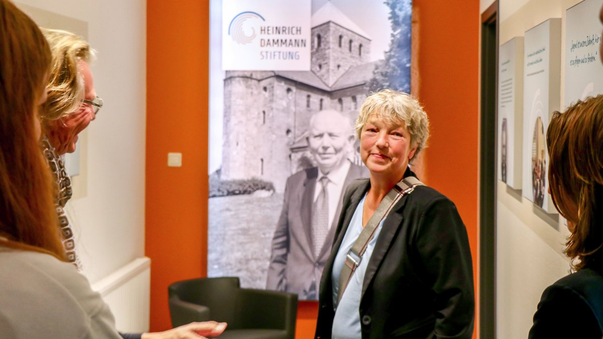Landtagspräsidentin Hanna Naber in den Räumen der Heinrich-Dammann-Stiftung. Hinter ihr ist eine orange gestrichene Wand zu sehen, an der ein beleuchtetes Schwarz-Weiß-Foto von Heinrich Dammann mit dem Logo der Stiftung hängt. Am Bildrand sind im Vordergrund mehrere Menschen der Stiftung zu erkennen.