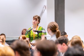Bild: © Niedersächsischer Landtag Zwei junge Frauen des jungen Europaparlamentes stehen während der Debatte an den Mikrofonen zwischen den Sitzreihen und sehen sich gegenseitig an. Um sie herum sitzen einige andere Schülerinnen und Schüler.