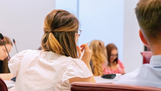 Mehrere junge Menschen sitzen an einem Konferenztisch. Im Fokus eine junge Frau mit einem Kugelschreiber in der Hand, die aufmerksam zuzuhören scheint.