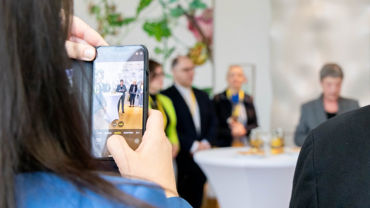 Im Fokus ein Handy, mit dem gerade ein Foto von Präsidentin Naber beim Sprechen gemacht wird. Im Hintergrund unscharf das Fotomotiv.