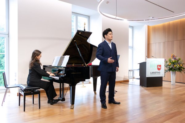 Bild: © Niedersächsischer Landtag Ein Mann im eleganten blauen Anzug steht neben einem Flügel, während eine Frau am Klavier sitzt und Notenblätter auf dem Instrument liegen. Der Raum hat hölzernen Boden und große Fenster im Hintergrund.
