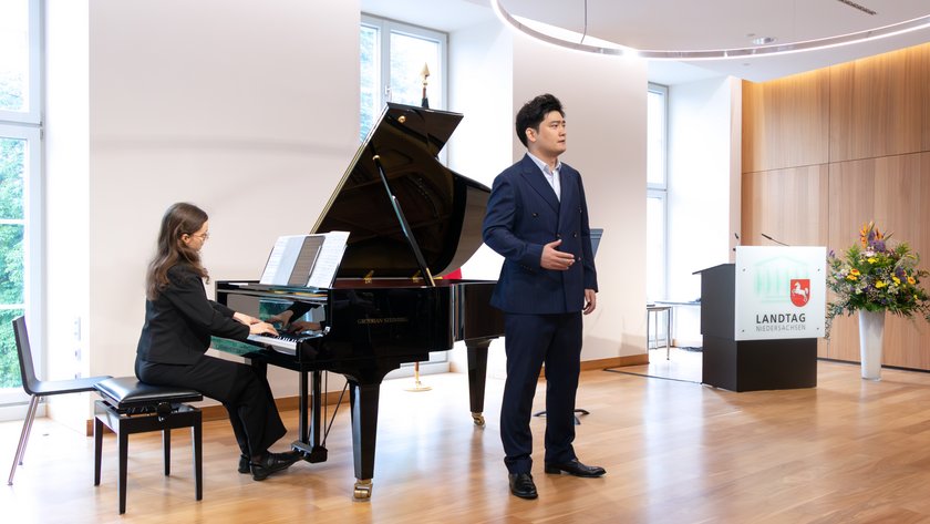 Ein Mann im eleganten blauen Anzug steht neben einem Flügel, während eine Frau am Klavier sitzt und Notenblätter auf dem Instrument liegen. Der Raum hat hölzernen Boden und große Fenster im Hintergrund.