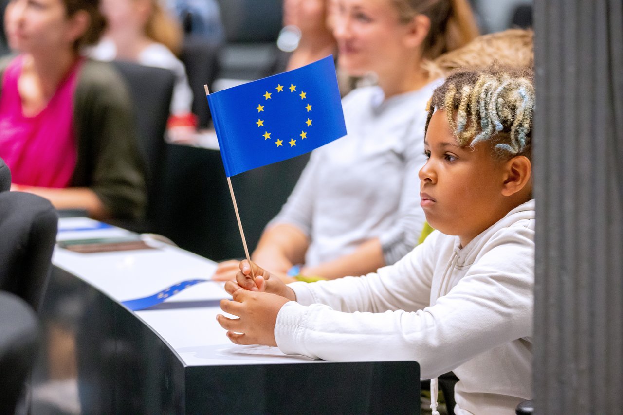 Ein Schüler sitzt im Forum des Landtages und hält eine kleine Europaflagge, im Hintergrund weitere Schülerinnen und Schüler.