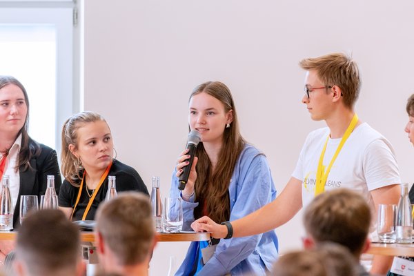 Klick auf das Bild öffnet eine vergrößerte Ansicht in einem Overlay. Zum Schließen des Overlays die ESC-Taste drücken. Vier Jugendliche während der Abschlussdiskussion, eine von ihnen spricht ins Mikrofon.'}