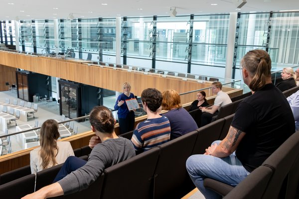 Bild: © Niedersächsischer Landtag Landtagslotsin Laura Koren steht vor einer Gruppe Menschen auf der Besuchertribüne im Plenarsaal und erklärt etwas anhand eines Schaubildes