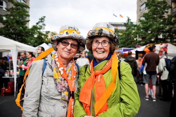 Bild: © Kirchentag/Jenna Dallwitz Zwei Damen mit bunten Hüten aus Zeitungen und orangen Schals lachen in die Kamera beim Kirchentag in Berlin 2017 (Abend der Begegnung)