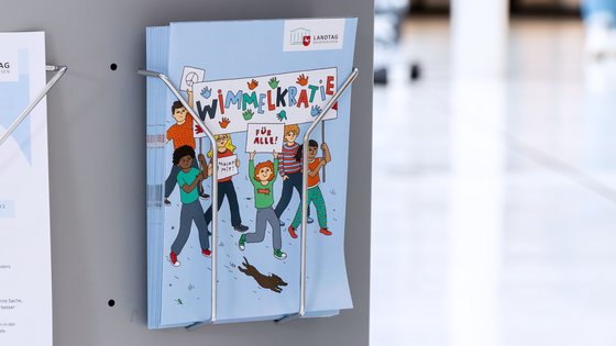 Flyer mit dem Titel "Wimmelkratie" in einem Flyerständer, auf dem Flyer sind bunte Illustrationen von Kindern zu sehen. Im Hintergrund unscharf die Portikushalle des Landtages.