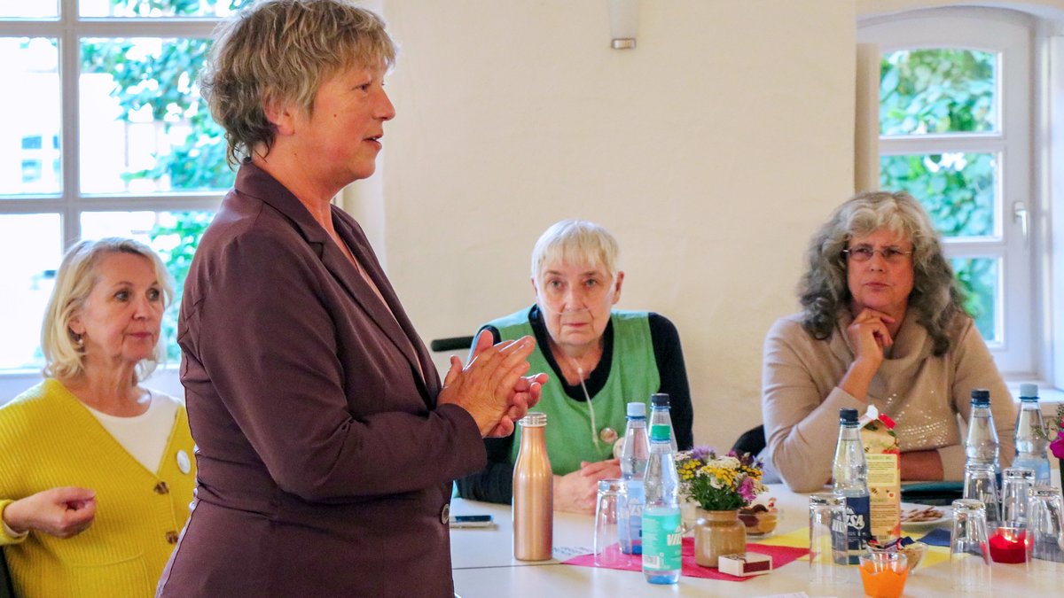Landtagspräsidentin Hanna Naber steht in einem Raum, im Hintergrund sitzen drei Frauen der Omas gegen Rechts an einem Tisch und hören ihr zu.