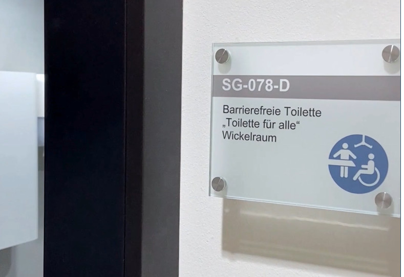 Hinweisschild vor dem Pflegebad mit dem Schriftzug "Toilette für alle" und Symbolen für die Barrierefreiheit.